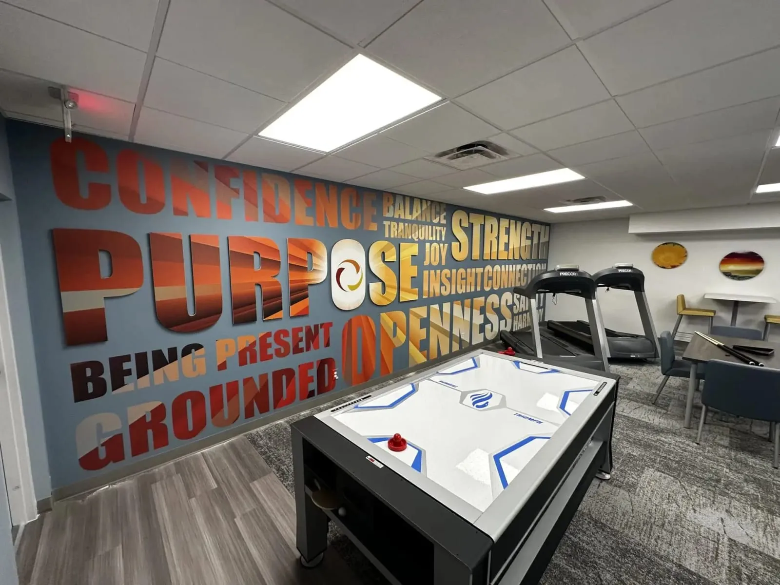 A lounge at Leora with a wall of words, Purpose, Confidence and Strength among them, an air hockey table and treadmills